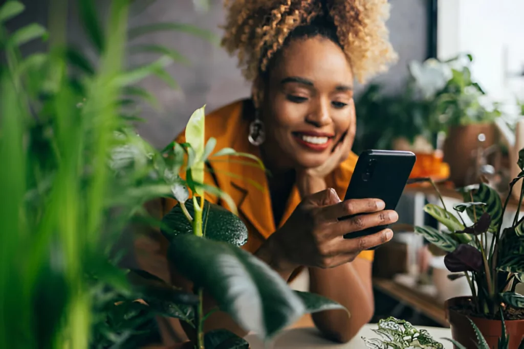 Uma mulher loira bonita feliz usando seu telefone celular enquanto cuida de suas plantas de casa