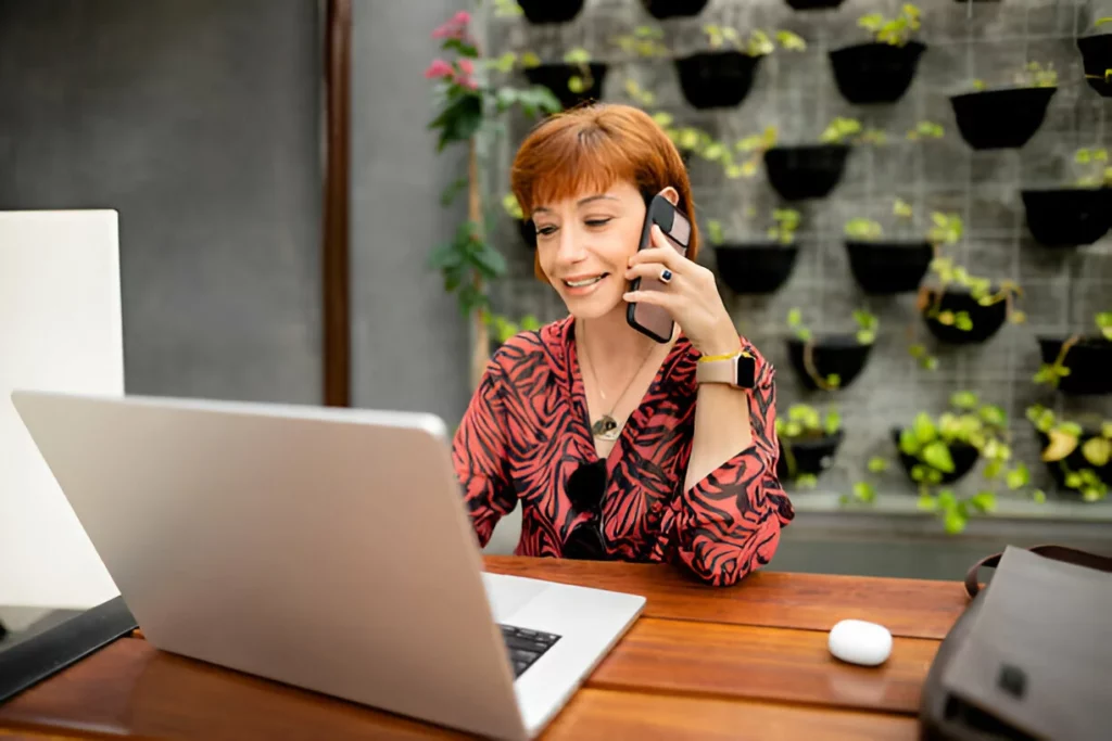 Mulher madura sorridente usando laptop com o FlowUp e falando ao telefone em um café